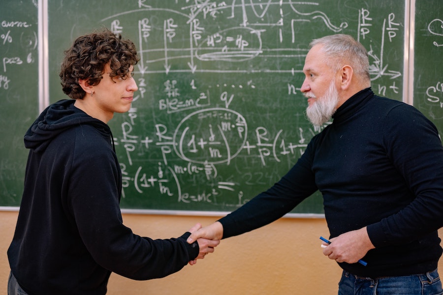 profesor preparador EVAU estrecha la mano de un alumno en un salón de clases frente a una pizarra